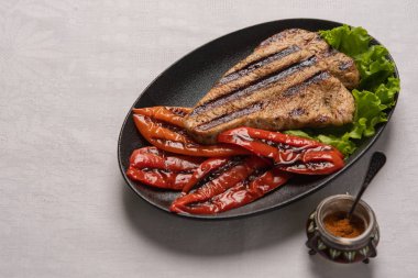 Pişmiş biber ve marul yaprakları ile ızgara Hindi göğüs biftek hafif keten arka plan üzerinde siyah bir tabakta. Kırmızı biberli biberli shaker'ın yanında. Yatay yönlendirme, kopyalama alanı