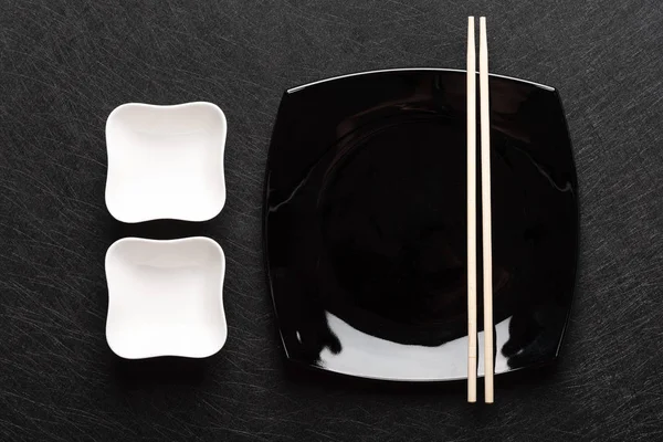 Çubuklar ve koyu bir arka plan üzerinde iki beyaz sos tekneler ile boş siyah plaka. Japon yemek tarzı. Üstte görüntü 