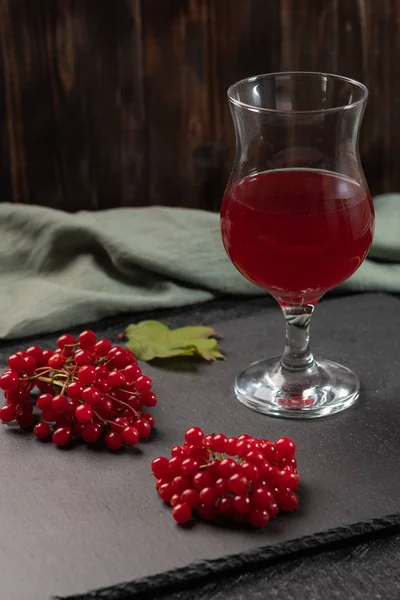 Red juice from viburnum in a glass on a stem on a dark background. Near viburnum berries and linen napkin. Healthy food