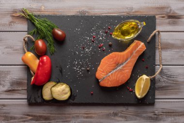 Çiğ somon bifteği. Karanlık bir tahtada. Pişmiş sebzelere, domateslere, limona yakın. Bir yağ da zeytinyağı. Sağlıklı yiyecekler. Üstte görüntü