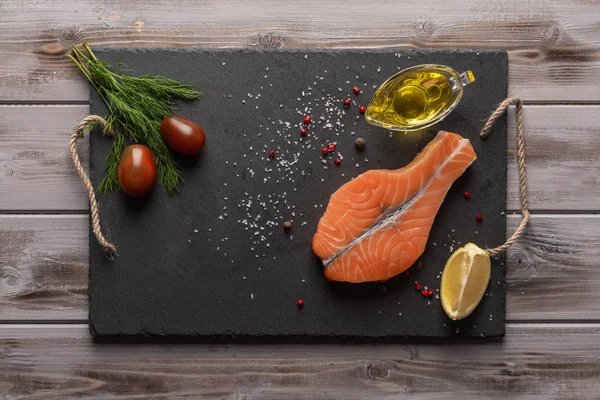 Çiğ somon bifteği. Karanlık bir tahtada. Domatesin, limonun yanında. Bir yağ da zeytinyağı. Sağlıklı yiyecekler. Üstte görüntü