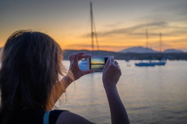 kadın arka görüş onu cep telefonu ile gün batımı bir fotoğraf çeker. Akıllı telefon ekranında çekim çerçevesini görebilirsiniz