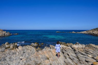 Mavi bir elbise kadın arka görünümü kayalık bir sahilde duruyor ve açık bir günde denize bakar
