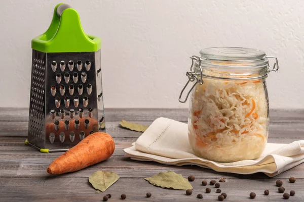 Chou en conserve fait maison dans un pot avec des feuilles de laurier sur une table en bois. Près de la râpe et des carottes. Style rustique — Photo