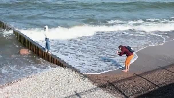 RÚSSIA, ZELENOGRADSK - 29 de setembro de 2020: Homem turista tira fotos em um smartphone em pé em uma menina quebra-mar contra o fundo de ondas com espuma — Vídeo de Stock