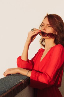 Kırmızı bir elbise ve güneş gözlüğü gün batımında balkonda genç kız esmer ayakta