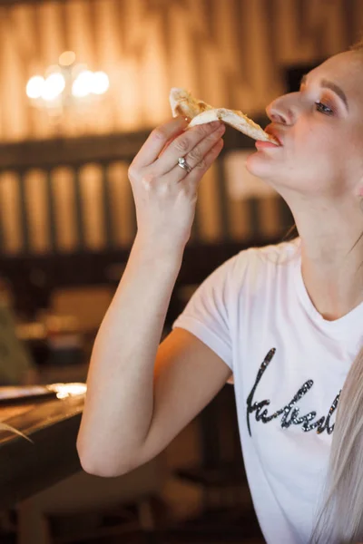 Casual girl eating pizza in the restaurant and enjoying this. Funny blonde girl in white t-shirt eating pizza at restaurant