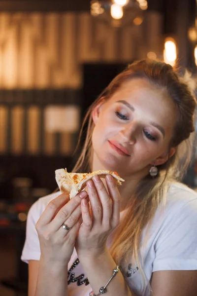 Casual girl eating pizza in the restaurant and enjoying this. Funny blonde girl in white t-shirt eating pizza at restaurant