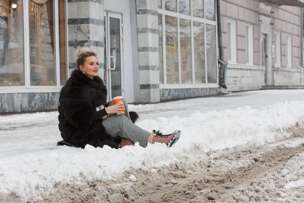 girl sitting on a snow in the city on curb covered with snow. Lifestyle. winter mood, coffee cup, mink fur clothing.