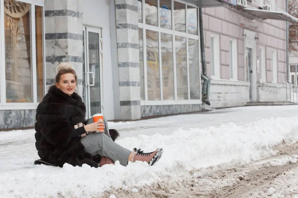 girl sitting on a snow in the city on curb covered with snow. Lifestyle. winter mood, coffee cup, mink fur clothing.