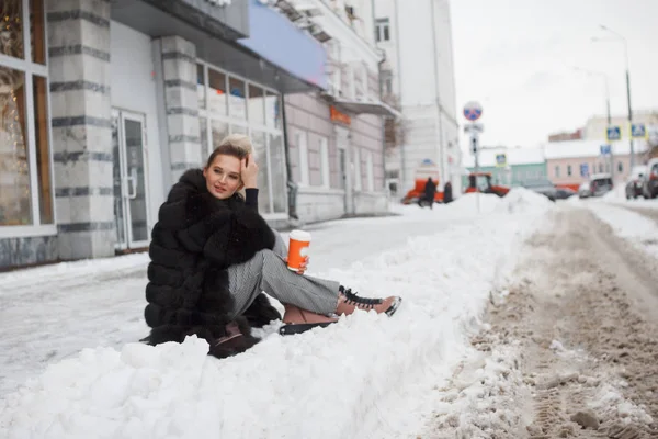 girl sitting on a snow in the city on curb covered with snow. Lifestyle. winter mood, coffee cup, mink fur clothing.