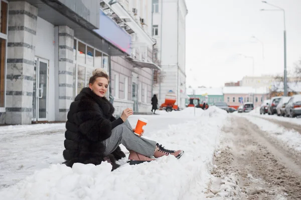 girl sitting on a snow in the city on curb covered with snow. Lifestyle. winter mood, coffee cup, mink fur clothing.