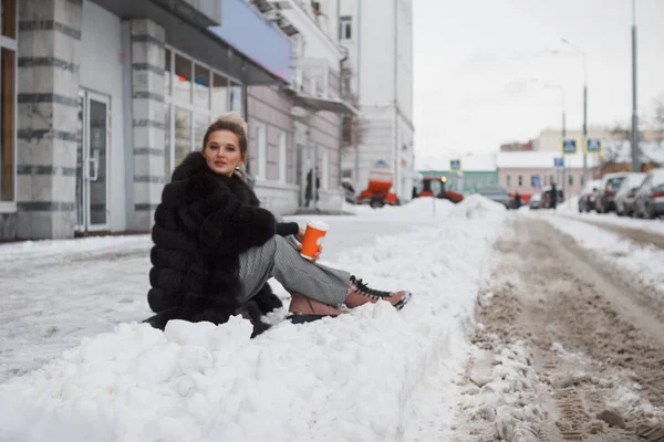 girl sitting on a snow in the city on curb covered with snow. Lifestyle. winter mood, coffee cup, mink fur clothing.