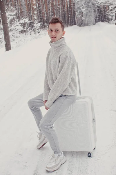 Portrait of young fashionable man in white clothes walking on snow holding suitcase and go pathway pine forest. Winter vacation travel concept. Outdoors in winter. Ski resort, mounting skiing resort