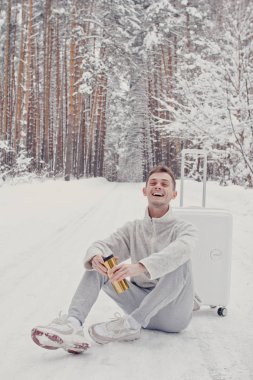 Bavul ve gitmek yolu çam ormanı holding kar üzerinde oturan beyaz giysili genç moda adam portresi. Kış tatil seyahat kavramı. Kışın açık havada. Kayak Merkezi, Kayak resort montaj