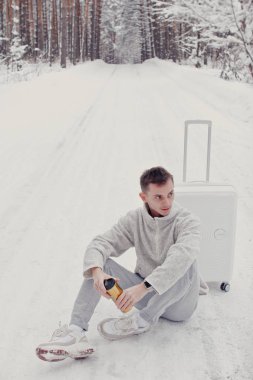 Bavul ve gitmek yolu çam ormanı holding kar üzerinde oturan beyaz giysili genç moda adam portresi. Kış tatil seyahat kavramı. Kışın açık havada. Kayak Merkezi, Kayak resort montaj