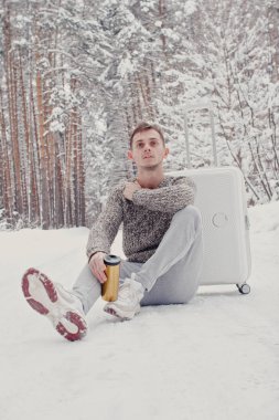 Bavul ve gitmek yolu çam ormanı holding kar üzerinde oturan beyaz giysili genç moda adam portresi. Kış tatil seyahat kavramı. Kışın açık havada. Kayak Merkezi, Kayak resort montaj