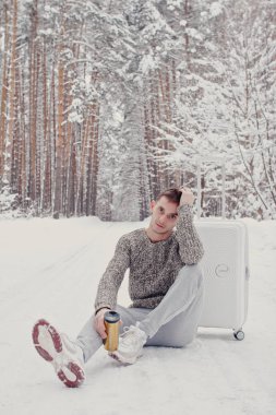 Bavul ve gitmek yolu çam ormanı holding kar üzerinde oturan beyaz giysili genç moda adam portresi. Kış tatil seyahat kavramı. Kışın açık havada. Kayak Merkezi, Kayak resort montaj
