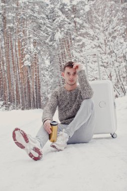 Bavul ve gitmek yolu çam ormanı holding kar üzerinde oturan beyaz giysili genç moda adam portresi. Kış tatil seyahat kavramı. Kışın açık havada. Kayak Merkezi, Kayak resort montaj