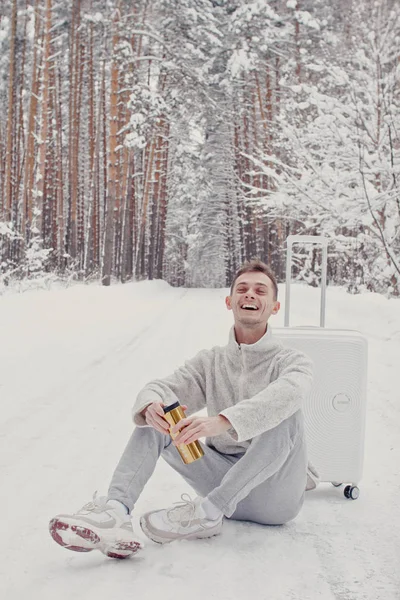 Bavul ve gitmek yolu çam ormanı holding kar üzerinde oturan beyaz giysili genç moda adam portresi. Kış tatil seyahat kavramı. Kışın açık havada. Kayak Merkezi, Kayak resort montaj
