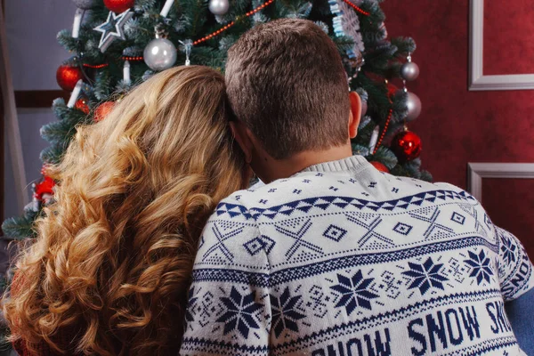 Couple in love sitting next to a Christmas tree, wearing warm sweaters, hugging and looking away from the camera towards the tree.