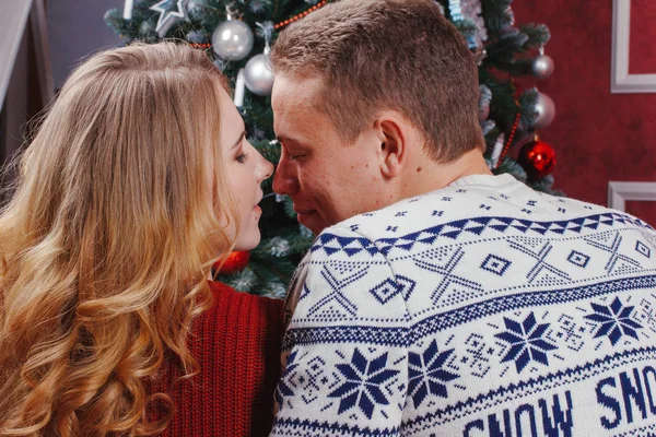 Couple in love sitting next to a Christmas tree, wearing warm sweaters, hugging and looking away from the camera towards the tree.