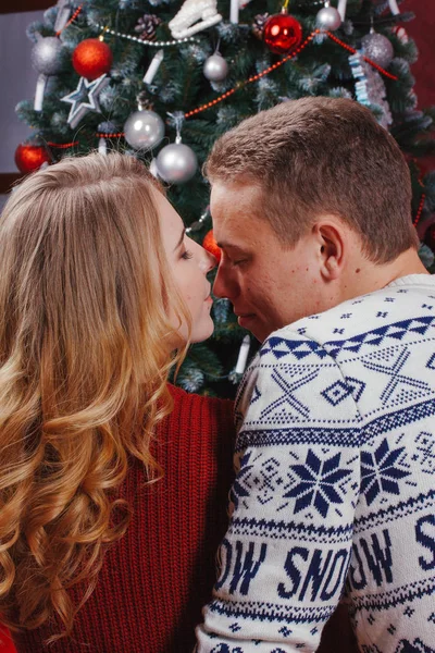 Couple in love sitting next to a Christmas tree, wearing warm sweaters, hugging and looking away from the camera towards the tree. — Stock Photo, Image