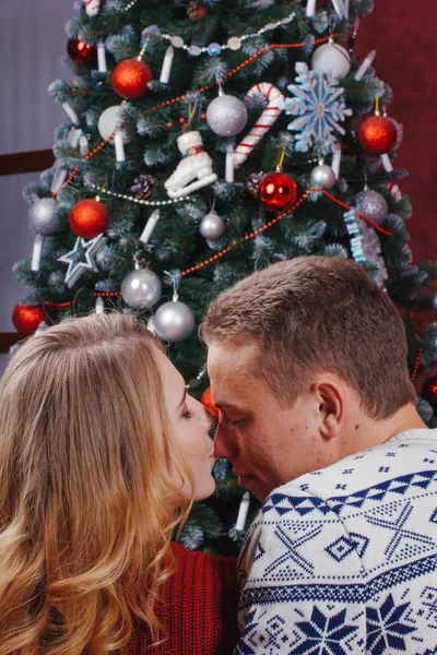 Dvojice zamilovaná, která sedí vedle vánočního stromu, nosí teplé svetry, objímá se a dívá se od kamery ke stromu. — Stock fotografie