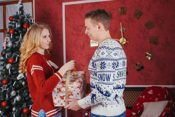 Des jeunes gens heureux se donnent mutuellement des cadeaux près de la cheminée près du sapin de Noël. Couple amoureux sont pleins d'anticipation pour la nouvelle année Image En Vente