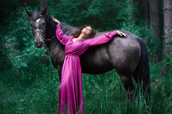 Portrét mladé krásné usmívající se brunetové ženy v růžovém hedvábném oděvu na tmavém koni v létě zelený Les. — Stock fotografie