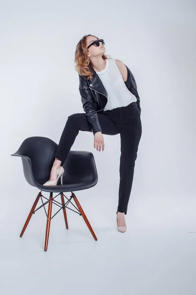 Pretty young fashion sensual woman posing on white wall background with black chair dressed in hipster style leather jacket and sunglasses outfit. Stylish fashionable girl hipster in black clothes.