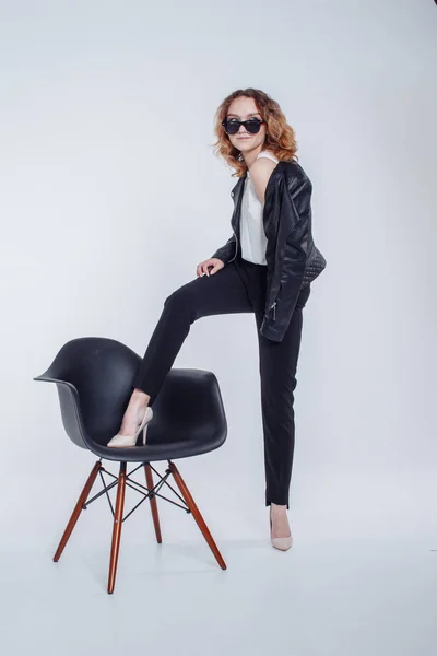 Mujer sensual de moda bastante joven posando sobre fondo de pared blanco con silla negra vestida con chaqueta de cuero estilo hipster y traje de gafas de sol. Elegante chica de moda hipster en ropa negra . —  Fotos de Stock
