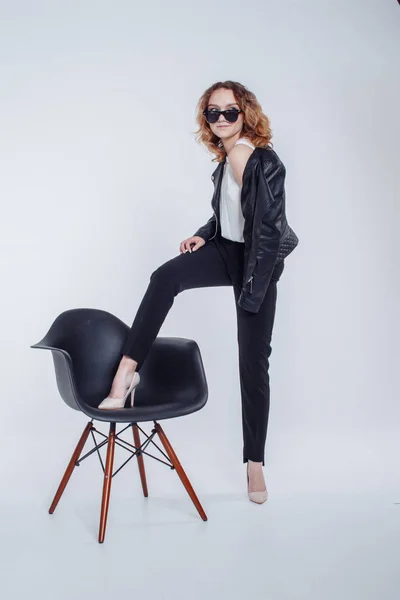 Pretty young fashion sensual woman posing on white wall background with black chair dressed in hipster style leather jacket and sunglasses outfit. Stylish fashionable girl hipster in black clothes.