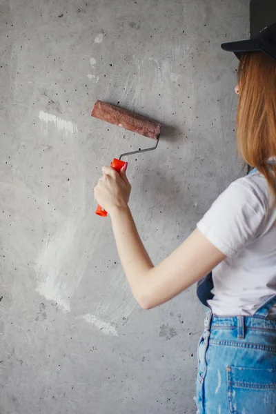 Enfoque selectivo en rodillo de pintura en la mano joven mujer caucásica delante de una nueva pared de reparación en una habitación . — Foto de Stock
