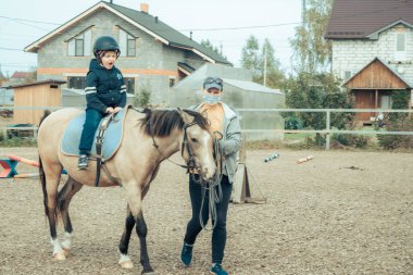 Turkuaz tulumlu küçük bir çocuk İzlanda midillisi atını komik bir perçemle okşuyor. Çocuk hipoterapiden sonra ata teşekkür ediyor. Eğitmen karantina sırasında maske kullanıyor.
