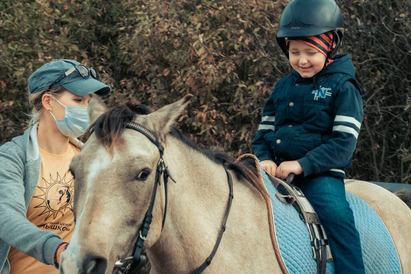 ターコイズ色の小さな男の子が アイスランドのポニーの馬を面白い前足で撫でています 子供はカバセラピーの後に馬に感謝します 講師は隔離中にマスクを使用します — ストック写真