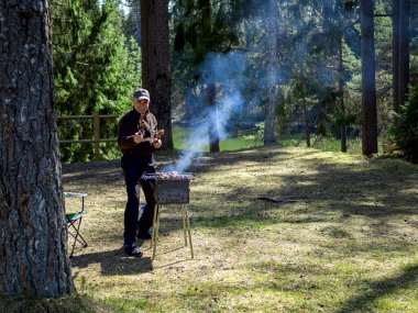 Yanan kömürün yanında ızgara yapmaya hazırlanan adam. Arka planda orman var. Barbekü ızgarası, barbekü.. 