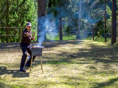 Yanan kömürün yanında ızgara yapmaya hazırlanan adam. Arka planda orman var. Barbekü ızgarası, barbekü.. 