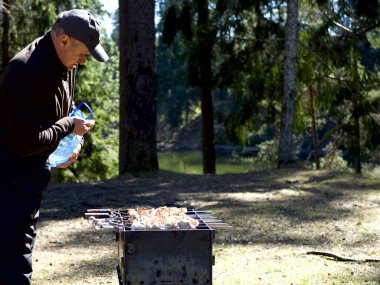 Yanan kömürün yanında ızgara yapmaya hazırlanan adam. Arka planda orman var. Barbekü ızgarası, barbekü.. 
