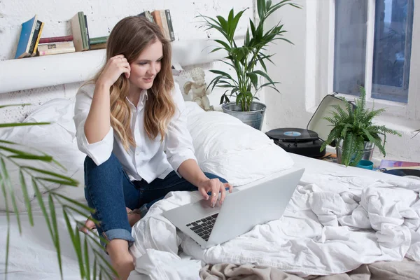 Schöne junge blonde Frau sitzt auf dem Bett mit Laptop, Jeans und weißem Hemd — Stockfoto