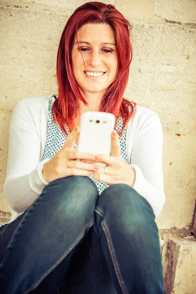 Улыбающаяся рыжая женщина, сидящая у старой стены в процессе отправки сообщений с мобильного телефона — стоковое фото