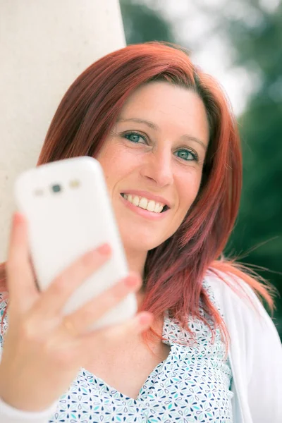 Cute smiling redheaded woman against an old wall with her mobile phone on the hand — Stock Photo, Image