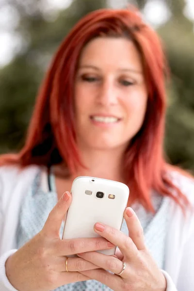 Улыбающаяся красивая молодая женщина вблизи мобильного телефона, на фоне летнего зеленого парка . — стоковое фото