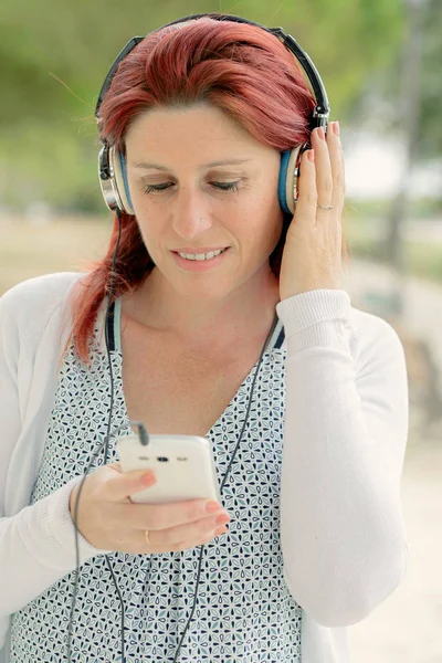 Atractiva pelirroja sonriente escuchando música en su teléfono con un auricular con vegetación en el fondo —  Fotos de Stock