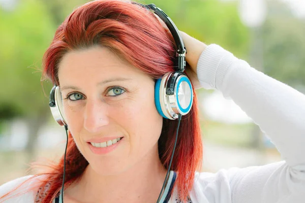 Attractive smiling redhead woman listening to music with greenery in the background — Stock Photo, Image