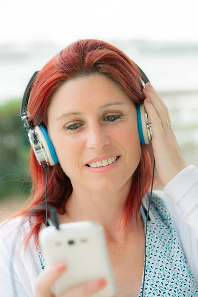 Attractive smiling redhead woman listening to music on her phone — Stock Photo, Image