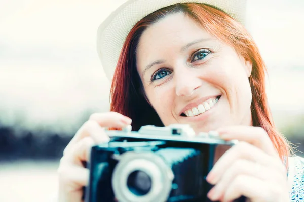 Retrato de una mujer pelirroja muy sonriente tomando fotos con una cámara vieja . — Foto de Stock