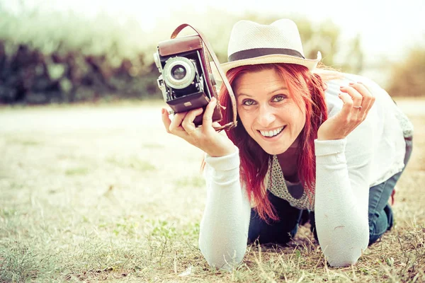 Pretty smiling redhead at ground taking pictures with an old camera. vintage treatment — Stock Photo, Image