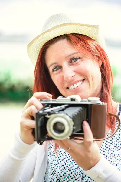 Portrait of a pretty smiling red-haired woman taking pictures with an old camera. — Stock Photo, Image