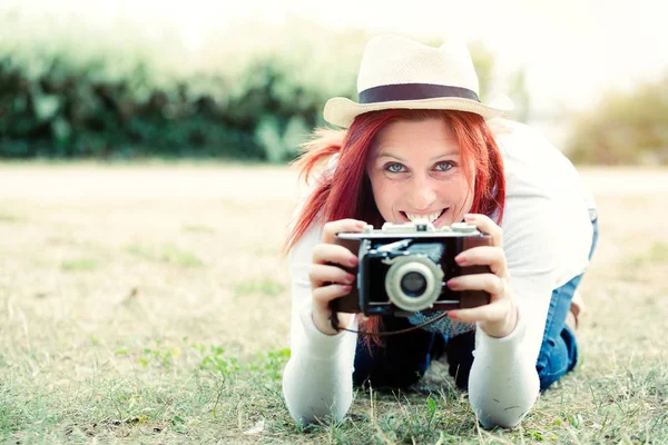 Pretty smiling redhead at ground taking pictures with an old camera. vintage treatment — Stock Photo, Image